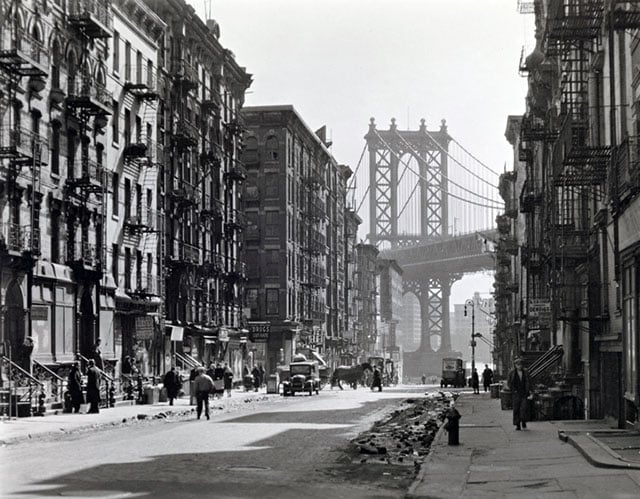 Pike and Henry Streets, Manhattan. Looking down Pike Street toward the Manhattan Bridge, street half in shadow, rubble in gutters, some traffic.