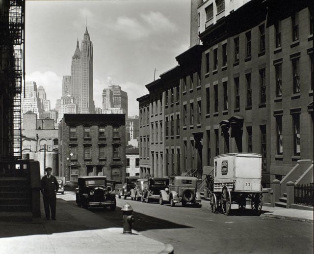 Willow and Poplar Street. Laundry wagon, cars, along sloping street lined with rowhouses, skyline of Manhattan visible above buildings at end of the street.