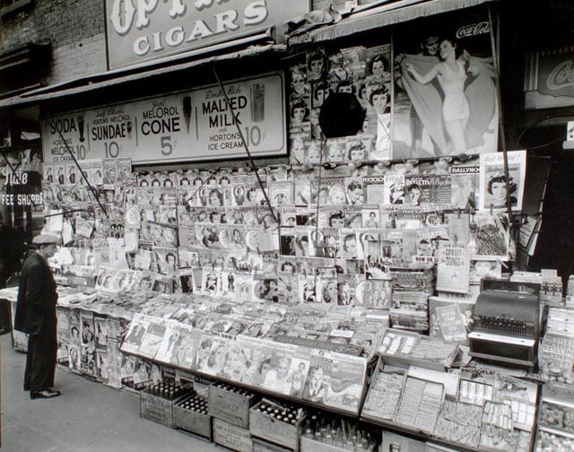 Newsstand, 32nd Street and Third Avenue, Manhattan. Newstand next to State Coffee Shoppe, large display of magazines, ads for sundaes, Coca-Cola above, boxes of sodas below, man at left