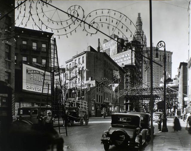 Oak and New Chambers Streets, Manhattan. Festive lights in curlicue designs arch over street, men with tall ladder, wagons, cars, billboards; 'el' and Municipal Bldg. just visible.