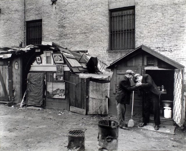 Huts and unemployed, West Houston and Mercer Street, Manhattan. Men share a light in front of hut with open door, milk can and washtub inside, hut to left has pictures in frames adorning the outside of it.
