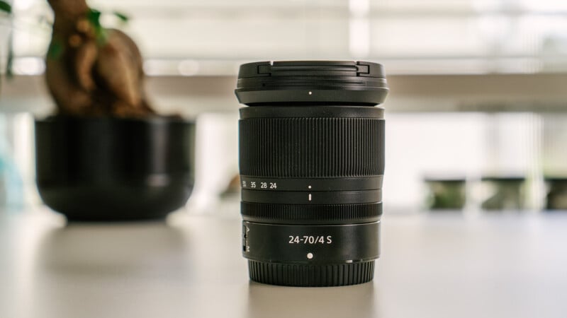 A Nikon 24-70mm lens sits on a white desk in front of a window.