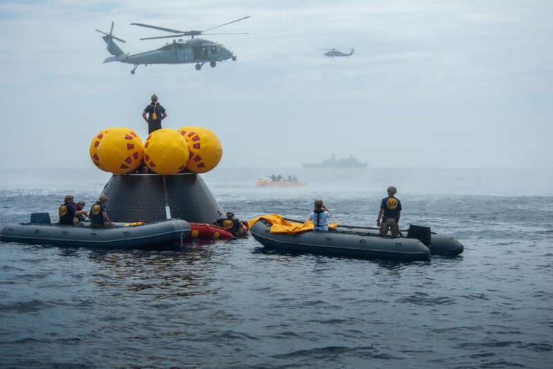 Naval personnel conduct a training exercise in the ocean with a submarine surfaced, surrounded by inflatable boats. helicopters hover above, and a distant ship is visible through the mist.