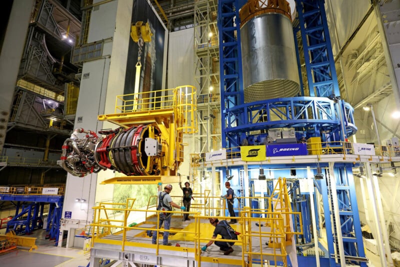 Engineers in a large industrial facility work on assembling a spacecraft with various tools and equipment, surrounded by yellow platforms and metallic structures.