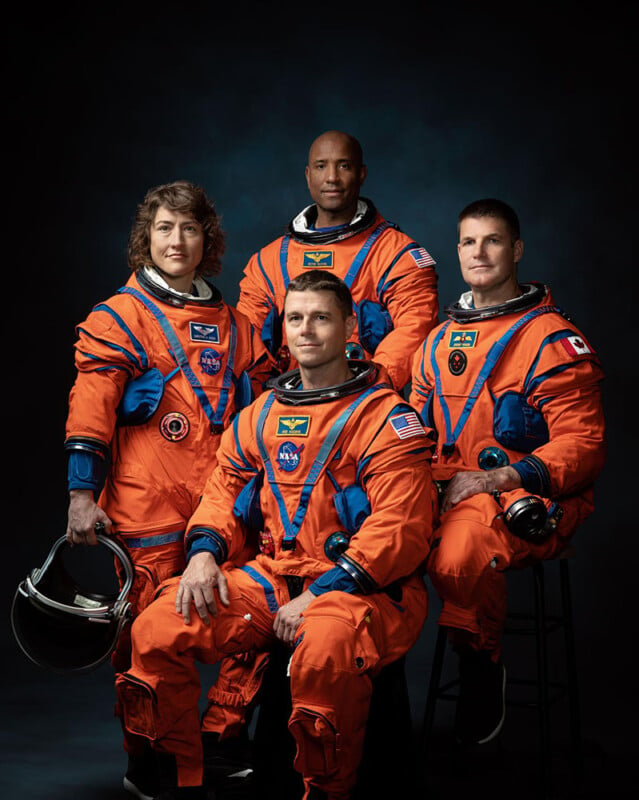 Four astronauts in orange nasa suits posing for a portrait against a dark background, three standing and one seated in front, holding a helmet.