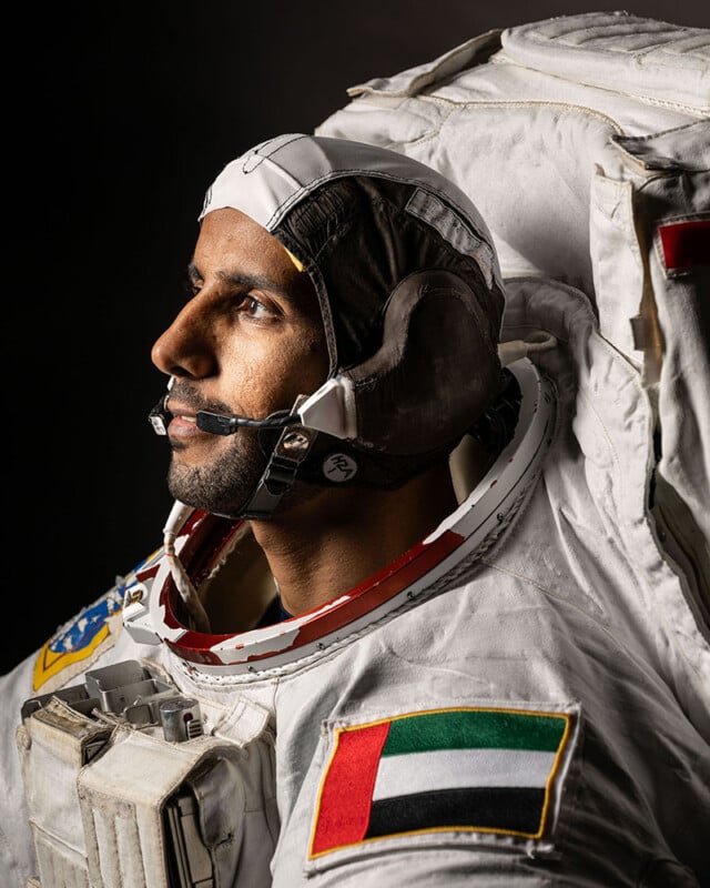 Profile portrait of an astronaut wearing a white space suit with a uae flag on the arm, looking thoughtfully to the side against a dark background.