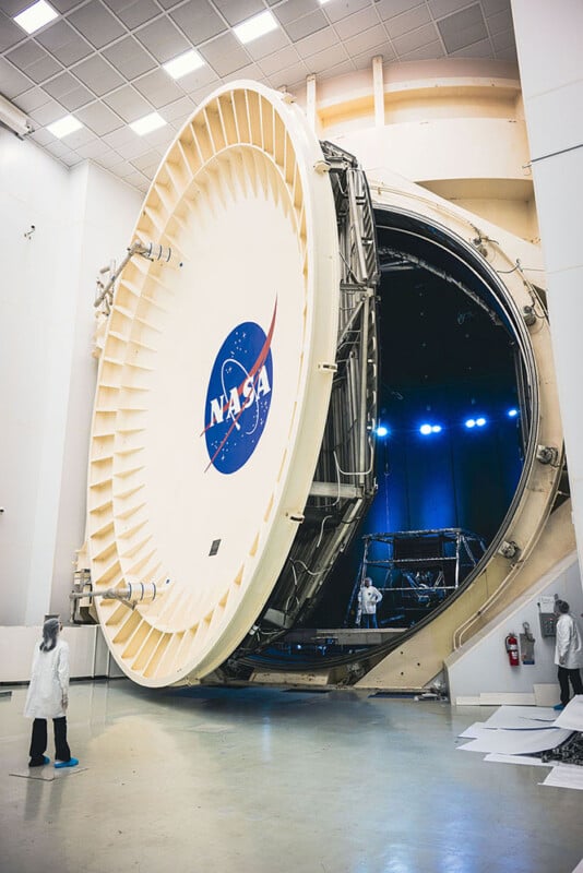 A large nasa vacuum chamber stands open in a facility, with engineers in lab coats observing and working around it. the chamber door features the nasa logo.
