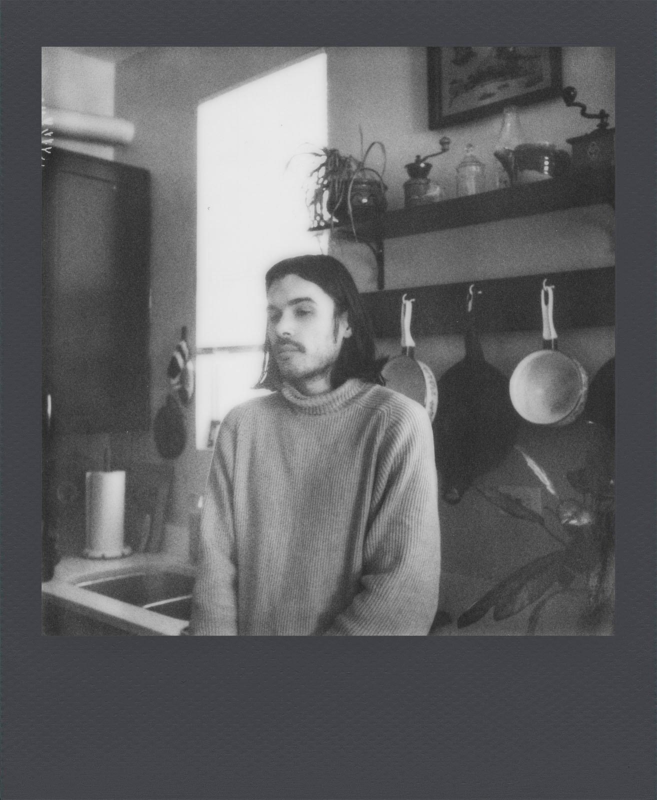 A black and white photo of a person with long hair and a beard, wearing a sweater, standing contemplatively in a cozy kitchen with pots and plants on shelves.