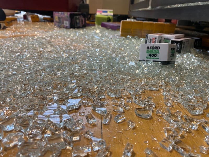 Broken glass scattered on the floor in a store aisle. Boxes of Ilford Delta 400 film and other films are visible in the background, along with various packages, suggesting the scene is in a photography or electronics store.