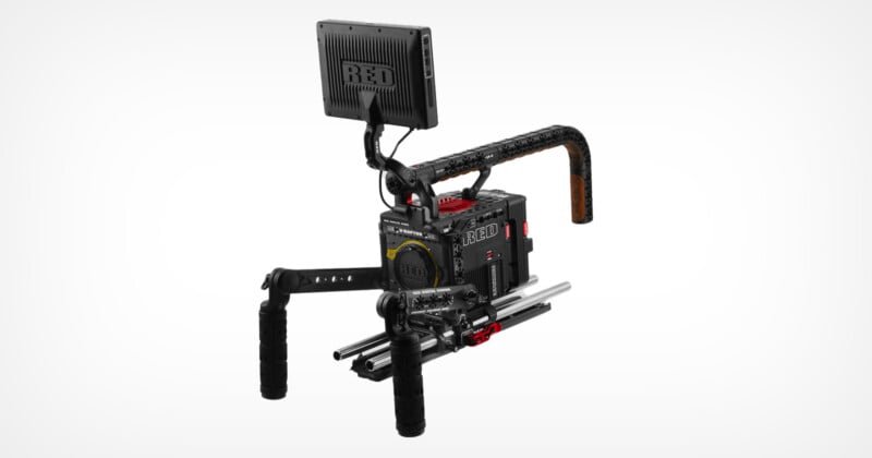 A professional cinema camera rig with a RED camera body, two hand grips, a top handle, a monitor mounted on top, and rods for accessories. The setup appears robust and is designed for filmmaking.