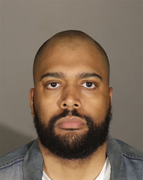 A person with a shaved head and a beard is looking directly at the camera. They are wearing a denim jacket over a white shirt. The background is plain and gray.