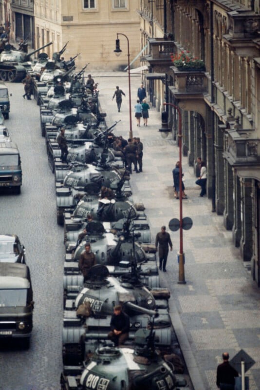 An aerial view of a cobblestone street filled with military tanks and soldiers. A few civilians walk along the sidewalk beside the buildings.