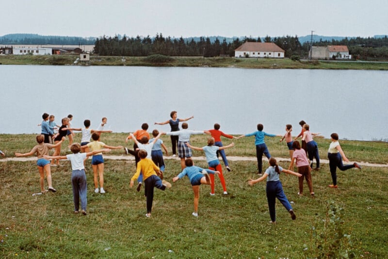 A group of people are doing exercises on a grassy area by a lake. They are stretching one leg back and extending their arms, while facing the water. There are buildings and trees in the background under a cloudy sky.