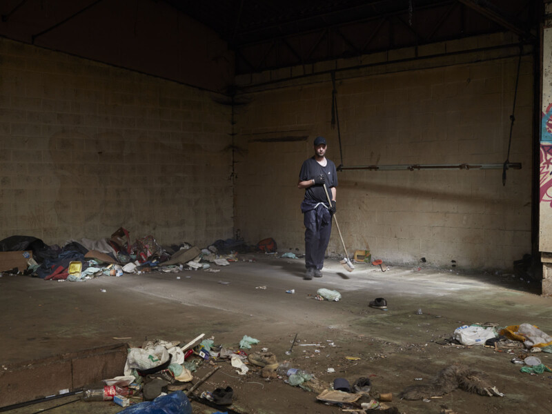 A person sweeps the floor of a dimly lit, abandoned building with scattered trash and debris. The walls are bare and dirty, and the overall scene appears neglected and rundown.