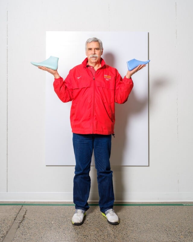 A person with gray hair and a mustache stands against a plain background, holding a blue shoe mold in each hand. They are wearing a red jacket, blue jeans, and white sneakers.