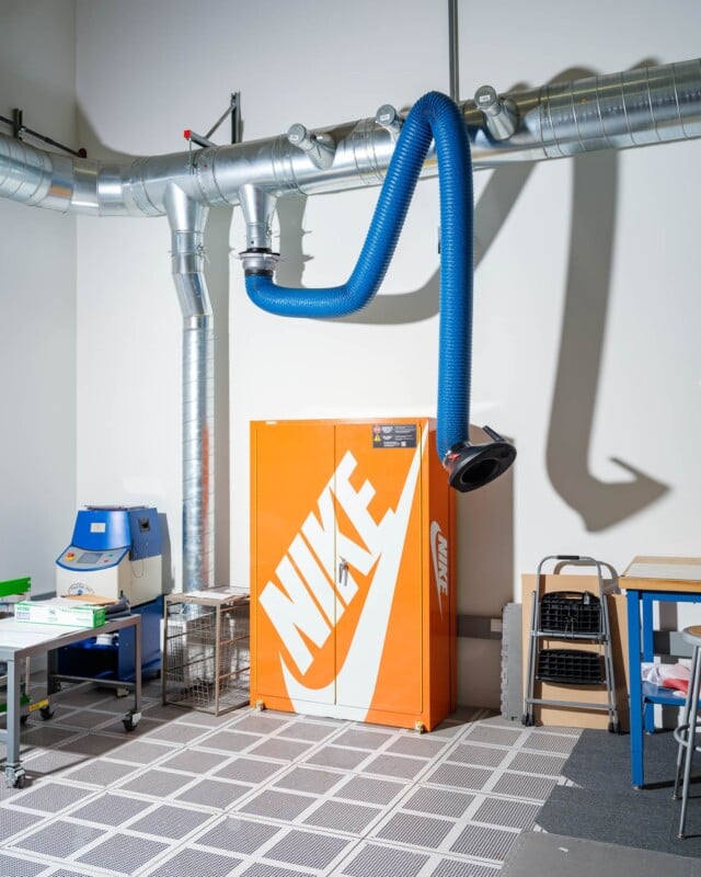 Industrial workspace featuring a prominent orange Nike cabinet with a large logo. Overhead, metallic ventilation pipes and a blue flexible duct are attached to the wall. Shelves and equipment are visible on the sides, set on a gridded floor.