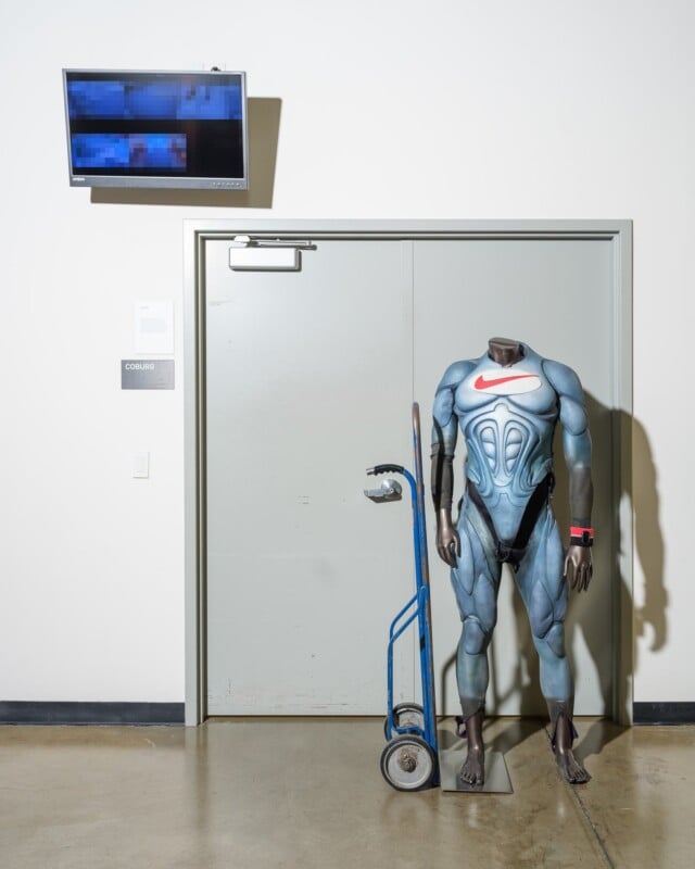 A headless mannequin wearing a futuristic, form-fitting suit with a red Nike logo stands next to a blue hand truck in front of a gray door. A small screen displays a blurred image above the door. The setting is industrial with a polished concrete floor.