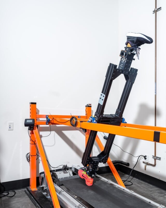 A robotic leg wearing sneakers is positioned on a treadmill setup. The apparatus is secured by an orange frame, with wires and a control box attached. The background wall is plain white with a visible electrical outlet and a pipe on the right.
