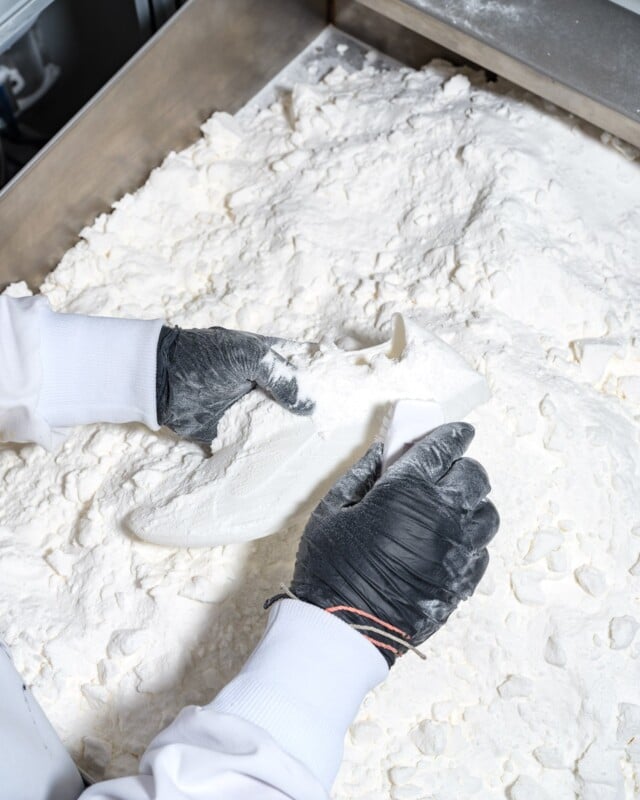 Gloved hands hold a shoe covered in white powdery material, surrounded by a large amount of similar powder on a surface, possibly in a manufacturing or artistic setting.