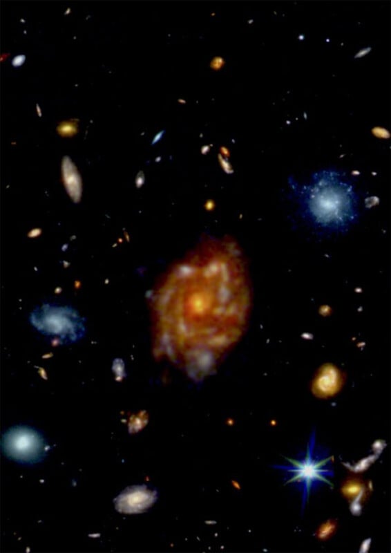 A vibrant image of deep space showing numerous galaxies. The scene includes spirals and ellipses of varying colors—blue, orange, red, and yellow—against a black backdrop. A bright star with diffraction spikes is visible at the bottom right.