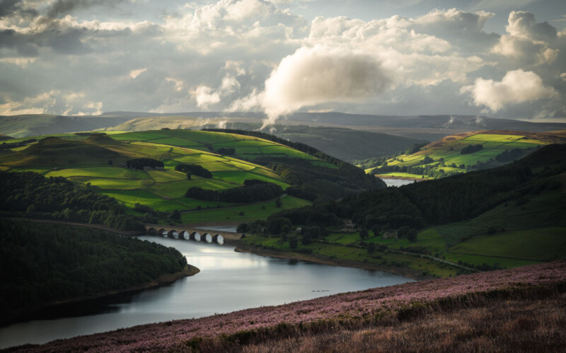 A picturesque landscape of rolling green hills with patches of sunlight, a river winding through the valley, and a stone bridge. Fluffy clouds scatter across the sky, casting soft shadows over the serene countryside.