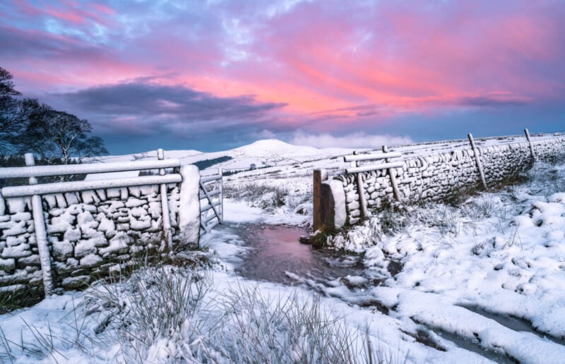 Snow-covered landscape with a stone wall and a metal gate leading to a snowy path. A mountain is in the distance under a vibrant pink and purple sky at sunset. Snow-laden grass and a small, icy stream are in the foreground.