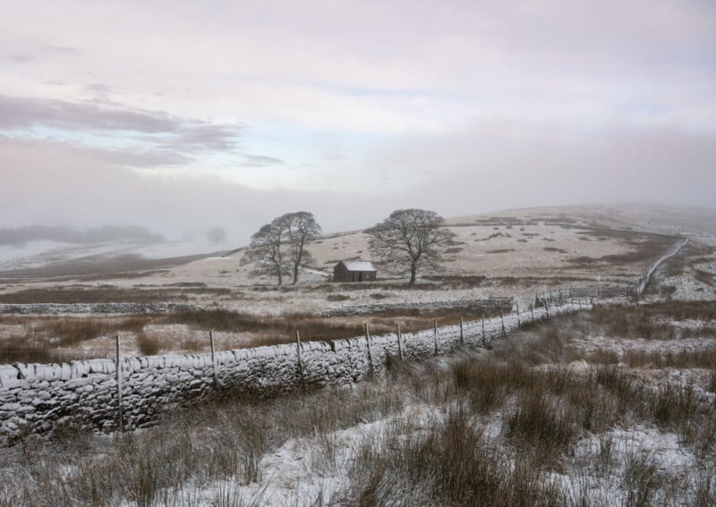 Snow-dusted landscape with a small stone building and two bare trees. A dry-stone wall runs through rolling fields under a cloudy sky, creating a serene winter scene.