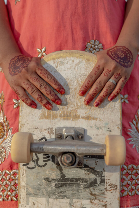 A person with intricate henna designs on their hands holds a worn skateboard vertically, against a pink outfit decorated with embroidered floral patterns.