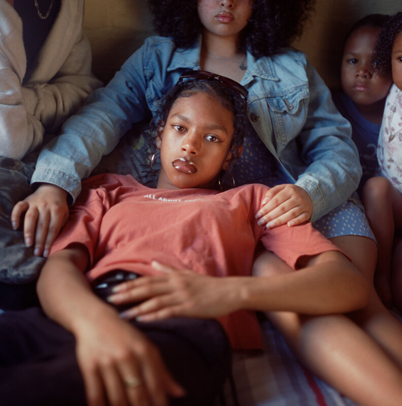 A young woman lies across the laps of others, looking thoughtfully at the camera. She wears a reddish shirt and hoop earrings, while those around her sit close together, conveying a sense of intimacy and support.