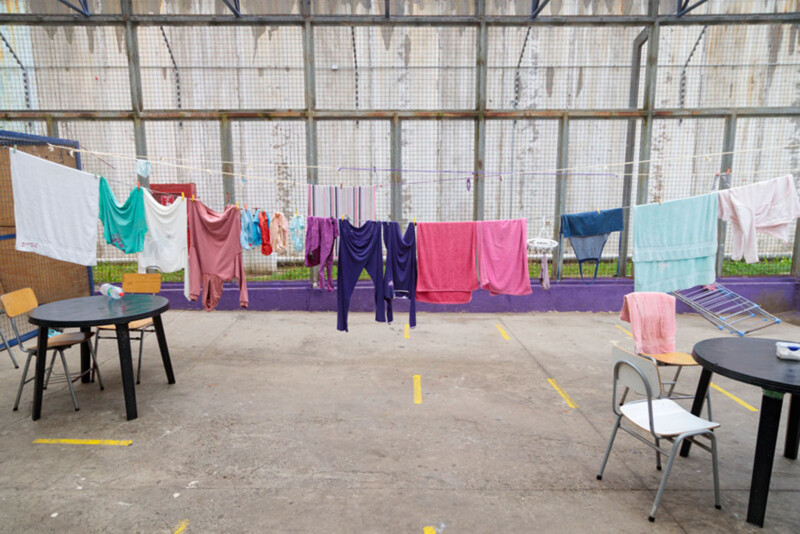 Clothes and towels of various colors hang on clotheslines in a fenced outdoor area with tables and chairs nearby. The ground is concrete, and a tall wall is visible in the background.
