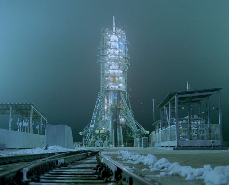 A rocket launch pad at night, illuminated by bright lights, stands surrounded by snow with train tracks leading toward it; the scene has a cold, teal-colored atmosphere.