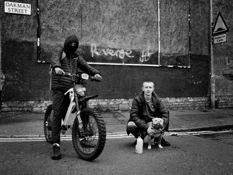 Two men pose on a street corner; one sits on a dirt bike wearing a mask, the other crouches beside a dog. A wall behind them has graffiti reading "Revenge," and a street sign says "Oakman Street.