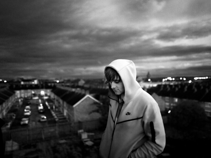 A young person wearing a hooded Nike jacket stands outdoors at dusk, looking down pensively. Rows of houses and parked cars stretch out behind them under a cloudy sky. The image is in black and white.