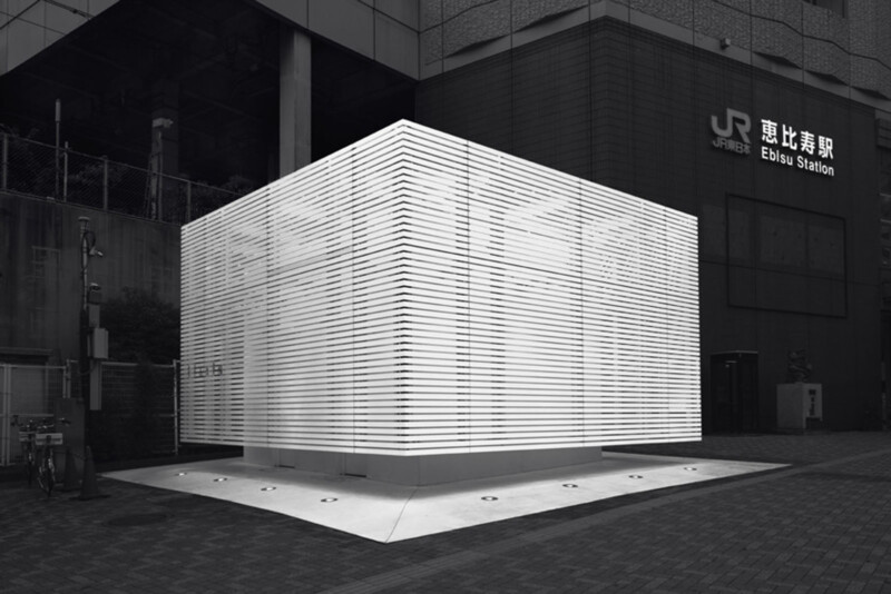 A brightly illuminated, modern public restroom with horizontal slats stands outside Ebisu Station in Tokyo at night, contrasting its glowing white structure against the dark urban background.