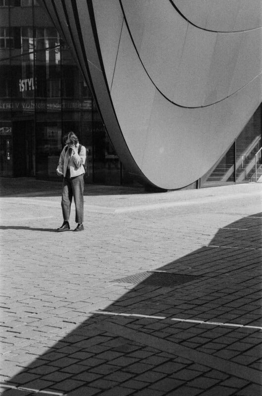 A person stands alone on a paved plaza near a modern building with large curved metal panels, partially in the shadow, while holding something to their ear, possibly a phone.