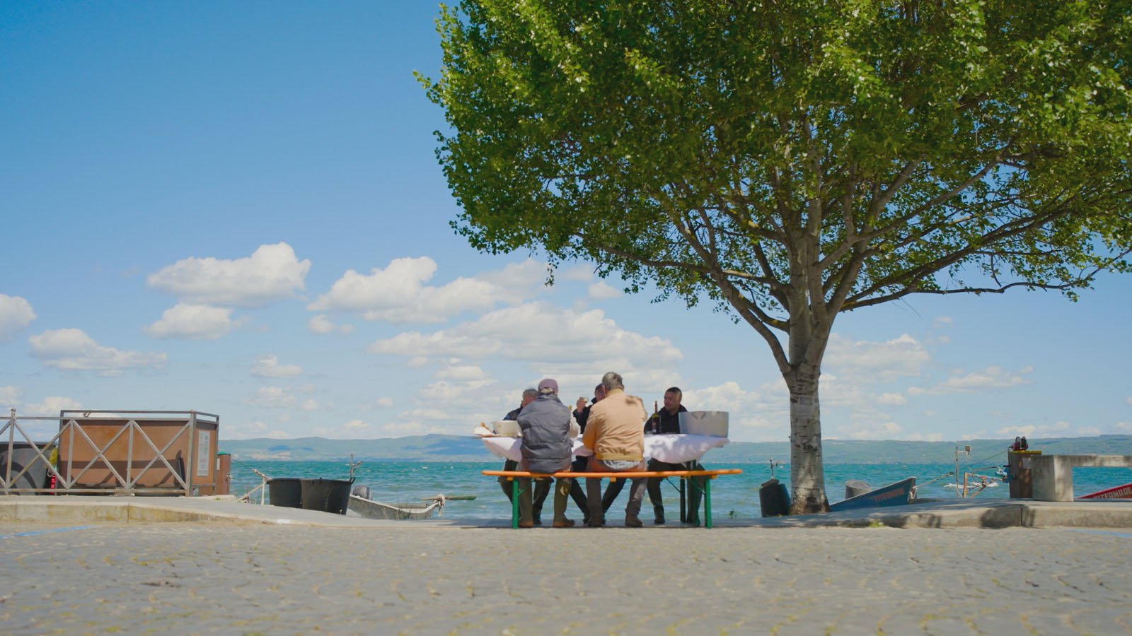 Three people sit at a table under a tree by the water, facing the lake on a sunny day with blue sky and scattered clouds. Boats and railings are visible nearby on the cobblestone ground.