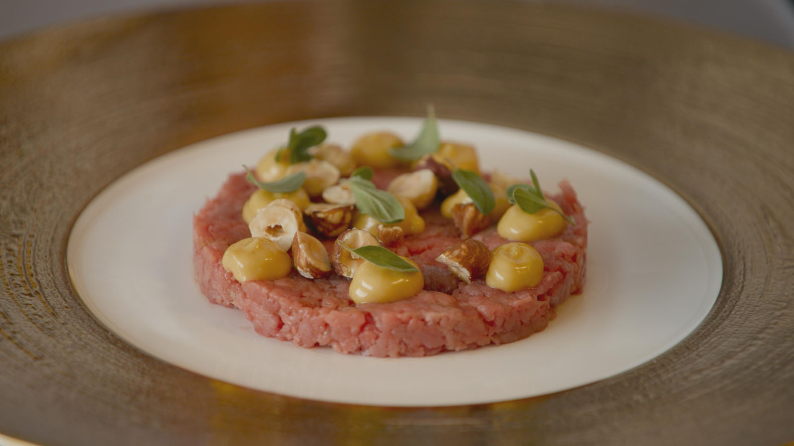 A round serving of beef tartare topped with dollops of yellow sauce, fresh herbs, and whole hazelnuts, presented on a white plate with a decorative gold rim.