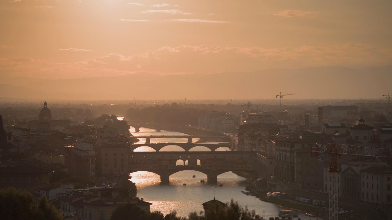 A sunset view of Florence, Italy, showing the Arno River with several historic bridges, including the Ponte Vecchio, and city buildings silhouetted against an orange sky.