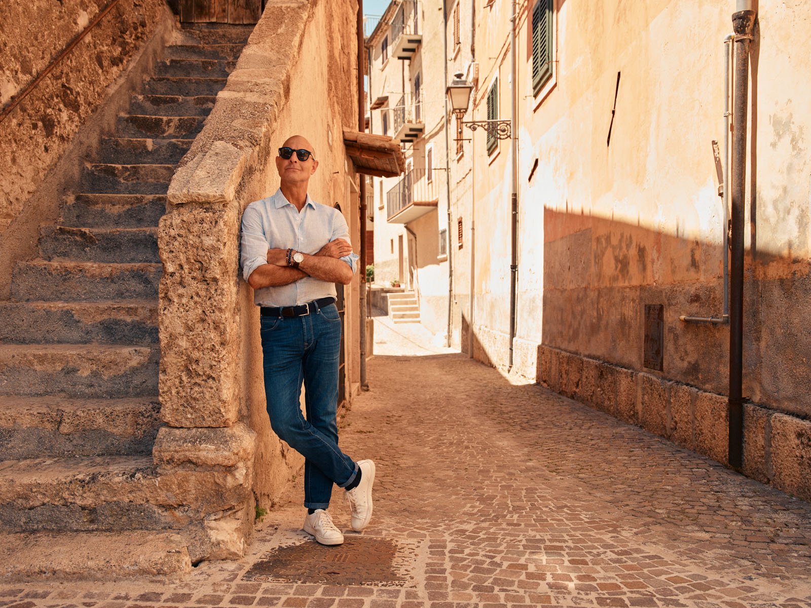 A man in sunglasses, jeans, and a light shirt stands with arms crossed, leaning against a stone staircase in a sunny, narrow, cobblestone street bordered by old buildings.
