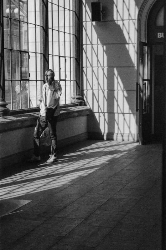 A person stands by large windows, talking on a phone. Sunlight streams through the window, casting strong shadows and grid patterns on the tiled floor and walls. The atmosphere is calm and contemplative.