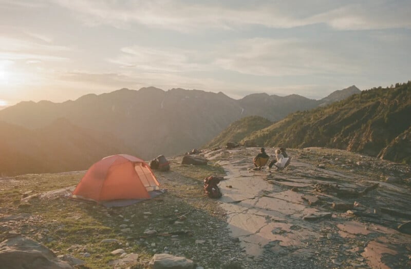 A red tent sits on rocky ground near a mountain edge at sunrise, with two people sitting nearby surrounded by backpacks and camping gear, overlooking distant mountain peaks.