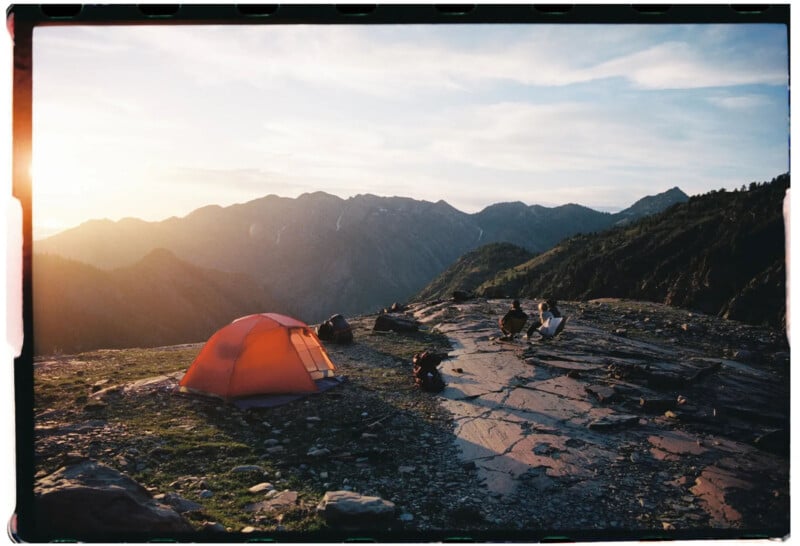 A bright orange tent is set up on a rocky mountain plateau at sunset, surrounded by camping gear and people near the edge, with mountain peaks in the background.