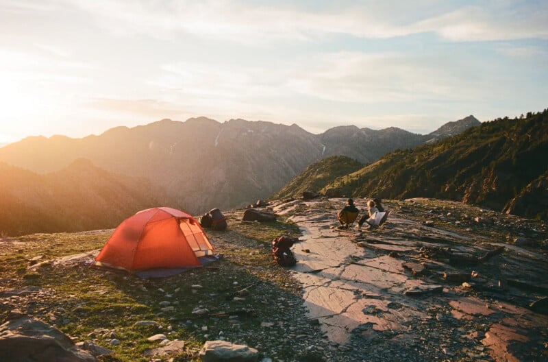 Two people sit on a rocky mountaintop near a red tent at sunset, surrounded by backpacks and camping gear, with mountain peaks and valleys in the background.