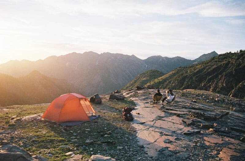 A bright orange tent sits on a rocky mountain plateau at sunset, with two people sitting nearby. Mountains with patches of snow rise in the background under a clear sky. Camping gear is scattered on the ground.