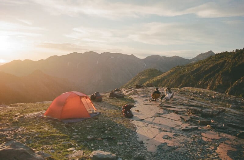 A red tent is pitched on rocky ground with gear scattered nearby. Two people sit facing a mountain range at sunset, with warm sunlight casting long shadows over the landscape.