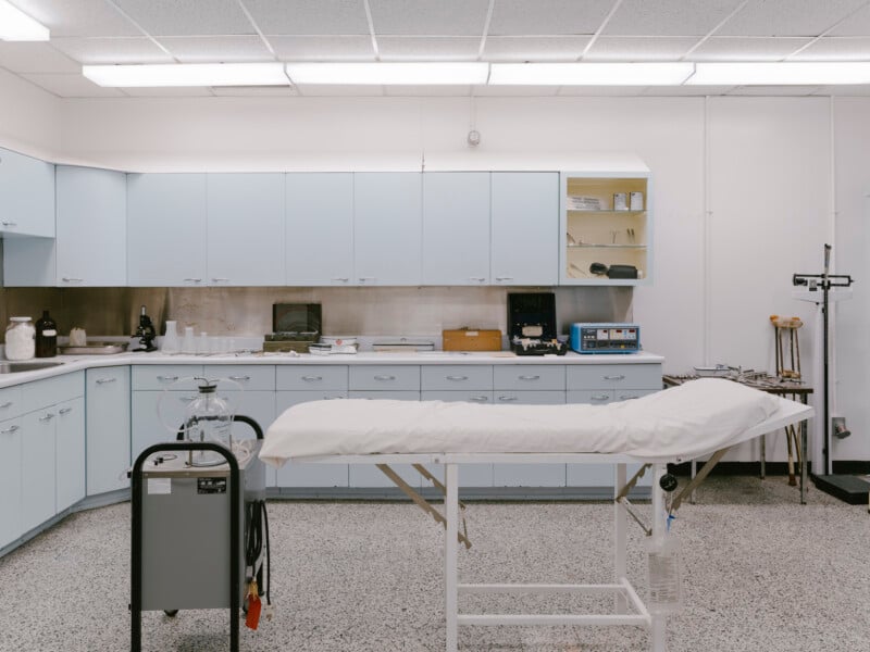 A clean, modern medical examination room with light blue cabinets, a white examination table, various medical equipment, and bright overhead lighting. The room has a sterile and organized appearance.