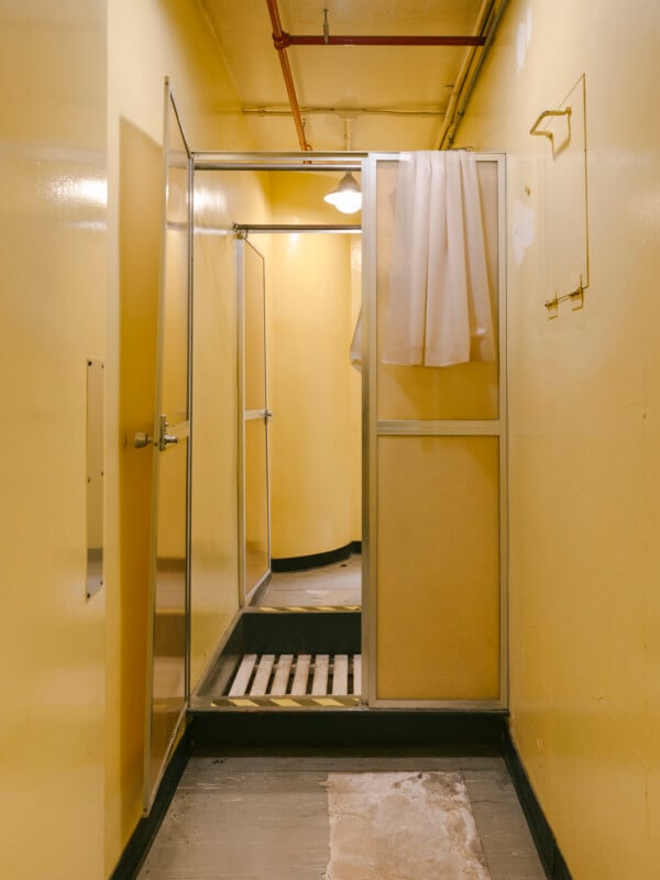 A narrow, yellow-painted room with an open glass shower stall, curtain partially hung, exposed pipes on the ceiling, and worn flooring. The lighting is dim, and the area appears old and utilitarian.