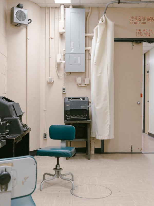 A vintage office room with beige walls, old electronic equipment, a blue swivel chair, and a partially drawn white curtain. There is a breaker box on the wall and French and English signs above a door.