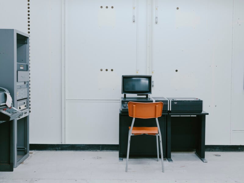 A vintage computer setup sits on a black desk with an orange chair in front, against a plain white wall. Old electronic equipment is visible on the left side of the image.