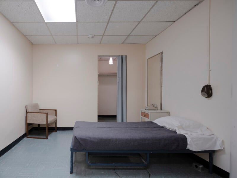 A sparse room with a single bed covered in gray bedding, a pillow, a small bedside table with a rotary phone, a wall lamp, an upholstered chair, and a closet with an open folding door under fluorescent lighting.